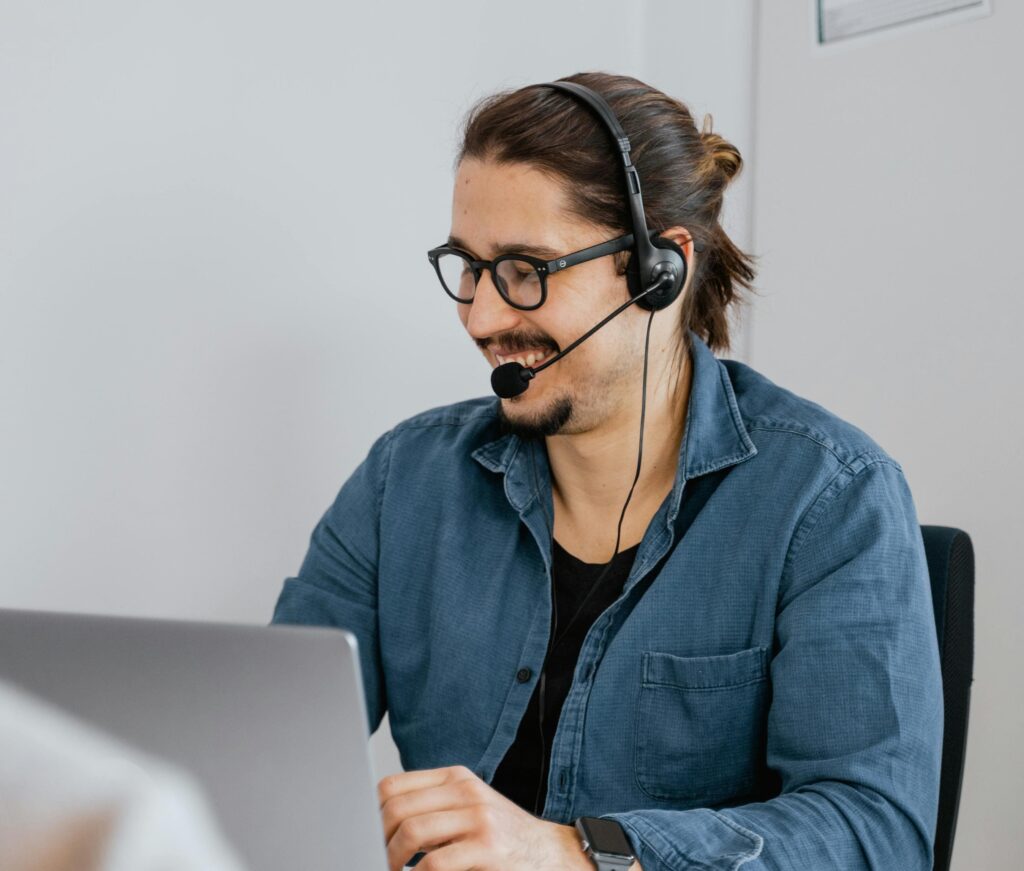 Ein lächelnder Mann winkt in einem Videoanruf von seinem Laptop aus, während er eine Tasse Kaffee hält und seine Kopfhörer um den Hals trägt, was die reibungslose Kommunikation mit Mitarbeiter im Ausland einstellen darstellt.